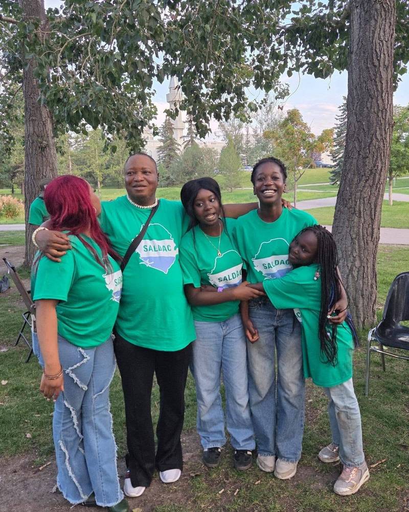 Five adults in green SALDAC T-shirts standing arm in arm under a tree at the 2026 picnic