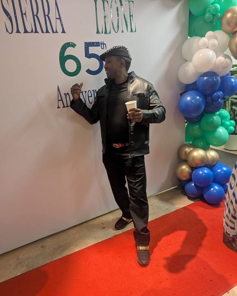 A guest in black posing on the red carpet in front of the Sierra Leone 65th anniversary backdrop and green, white and blue balloons